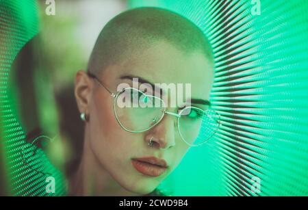 Image d'une belle jeune femme posant contre un panneau LED. Tête rasée à l'adolescence avec un aspect différent pour réaliser des portraits urbains Banque D'Images