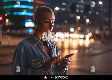 Image d'une jeune femme utilisant un téléphone portable dans la rue. Jeune fille aux cheveux rasés marchant le soir dans le centre-ville Banque D'Images