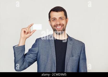 Jeune homme d'affaires avec une barbe dans une veste montrant une feuille de papier vierge. L'homme sourit et tient dans sa main montre une carte de visite. Portrait d'un homme sur fond gris. Banque D'Images