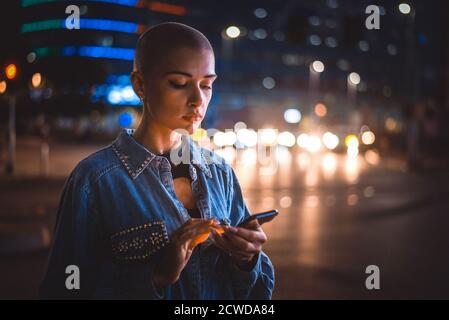 Image d'une jeune femme utilisant un téléphone portable dans la rue. Jeune fille aux cheveux rasés marchant le soir dans le centre-ville Banque D'Images