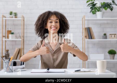 Moderne maison blogger, grand examen et passe-temps. Femme afro-américaine souriante montrant les pouces et regardant l'appareil photo, assis à une table avec un ordinateur portable Banque D'Images