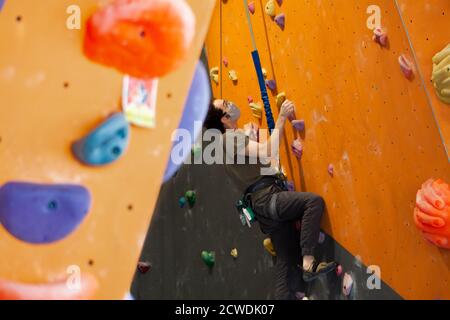 Un homme portant un masque pandémique COVID-19 ou coronavirus monte un mur attaché à une corde auto belay dans un gymnase d'escalade respecter les directives de distance sociale Banque D'Images