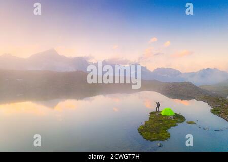 Randonneur avec tente buvant à partir d'une bouteille d'eau au lac Obere Schwarziseeli pendant un lever de soleil brumeux, col de Furka, canton d'Uri, Suisse Banque D'Images