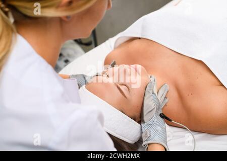 Le cosmétologue professionnel du matériel touche le visage du client avec des gants gris à microcourant pour la thérapie de soin de la peau de myostimulation, vue latérale Banque D'Images