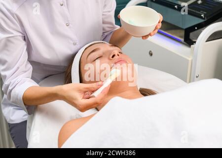 Les mains du cosmétologue tiennent un bol et une brosse cosmétique avec un masque facial bleu clair en argile à la boue pour les jeunes femmes, détendues sur un canapé, skincar professionnel Banque D'Images