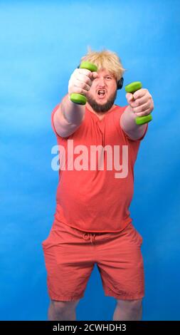 Drôle de chubby freak homme dans le casque et une perruque dans un t-shirt rose est engagé dans les haltères vertes et rires sur un fond bleu, vertical Banque D'Images