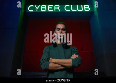 Portrait d'un joueur d'ordinateur dans un casque debout avec les bras croisés et regarder la caméra avec cyber club en arrière-plan Banque D'Images