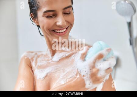 Jeune femme gaie utilisant le loofah exfoliant tout en prenant la douche Banque D'Images