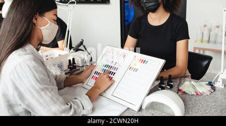 Nouvelle manucure après le verrouillage et choix de la couleur moderne après la quarantaine. La femme asiatique choisit le vernis à ongles dans le catalogue avec le maître afro-américain Banque D'Images