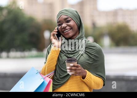 Femme musulmane noire avec sacs de shopping et café parlant Téléphone portable extérieur Banque D'Images