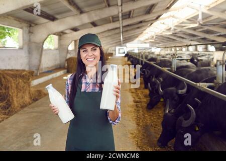 Bonne jeune femme debout dans un abri de vache, tenant des bouteilles de lait frais et regardant la caméra Banque D'Images
