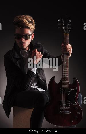 un guitariste sérieux portant des lunettes de soleil est assis et tient sa guitare électrique, sur fond noir studio Banque D'Images