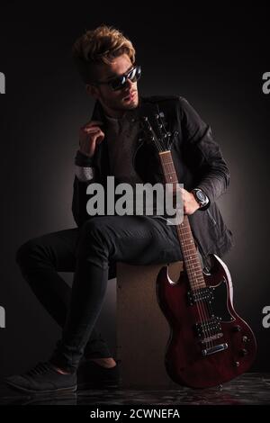 vue latérale d'un jeune guitariste assis et regardant en arrière pendant qu'il tient sa guitare électrique , sur fond de studio noir Banque D'Images