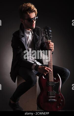 vue latérale d'un guitariste rock and roll tenant sa guitare électrique et pose pour la caméra en étant assis, sur fond noir studio Banque D'Images