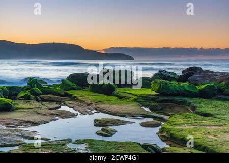 Paysage marin au lever du soleil avec des roches vertes de mousse à Killcare Beach sur la côte centrale, Nouvelle-Galles du Sud, Australie. Banque D'Images