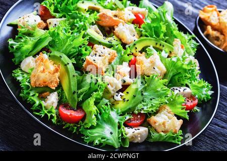 salade de feuilles avec avocat, tomates cerises, mozzarella et croûtons parsemés de graines de sésame noir sur une assiette, sur une table en bois, vue sur le paysage Banque D'Images