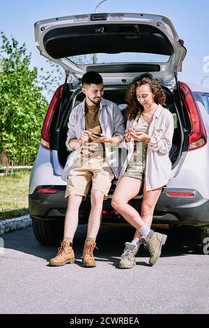 Jeunes randonneurs actifs positifs dans des bottes penchées sur le coffre de voiture et discuter de l'itinéraire de randonnée à l'aide des applications du téléphone Banque D'Images