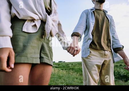 Gros plan d'un couple de voyage méconnaissable en chemises décontractées tenant les mains dans les collines en position debout dans le champ Banque D'Images