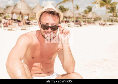 Jeune homme attrayant assis sur la plage, en train de retirer ses lunettes de soleil. Banque D'Images