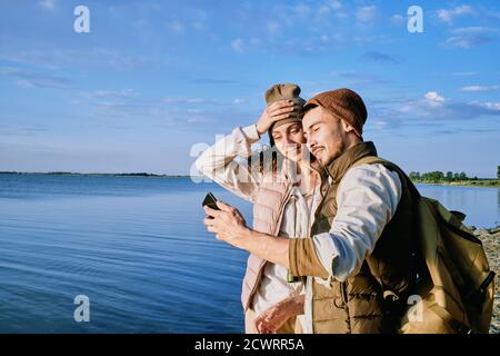 Contenu beau jeune couple en chapeaux et gilets debout à lac et prise de selfie sur smartphone pendant la randonnée Banque D'Images