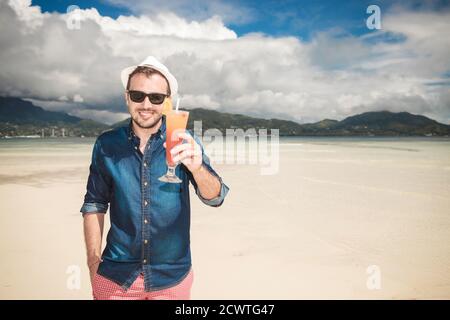 Joli jeune homme tenant une main dans sa poche tout en vous invitant à boire un cocktail froid, sur la plage. Banque D'Images