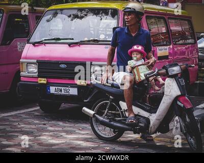 Un homme asiatique avec sa fille sur une moto tournée par le point de ramassage du colectivo sur l'île de Pangkor, en Malaisie Banque D'Images