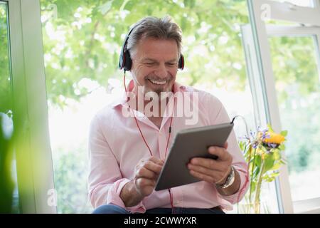 Homme senior heureux utilisant un casque et une tablette numérique à la fenêtre Banque D'Images
