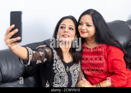 Deux amies souriantes assises sur un canapé à la maison et prenant un selfie avec le téléphone. Banque D'Images