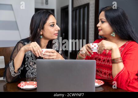 Deux amies assises à la maison avec un ordinateur portable devant elles, tenant des tasses à thé et parlant l'une à l'autre. Banque D'Images