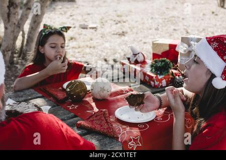 Deux filles célèbrent un pique-nique de Noël en plein air avec leur famille, portant des chapeaux de père Noël, avec une table décorée de cadeaux Banque D'Images