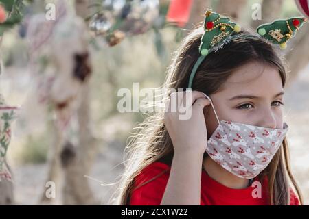 Une fille vêtue de rouge avec un bandeau de Noël, mettant sur un masque de protection médicale avec des dessins de Noël, dans la campagne Banque D'Images