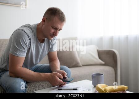 Homme adulte utilisant la calculatrice tout en étant assis sur le canapé et en gérant finances personnelles Banque D'Images