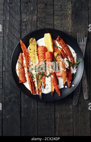 Salade de carottes rôties avec yaourt, épices et herbes sur fond sombre. Banque D'Images