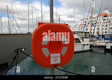 Utilisation en cas d'urgence uniquement. Une bouée Lifebuoy orange vif et un logement placé sur un poteau avec un fond de marina. Banque D'Images