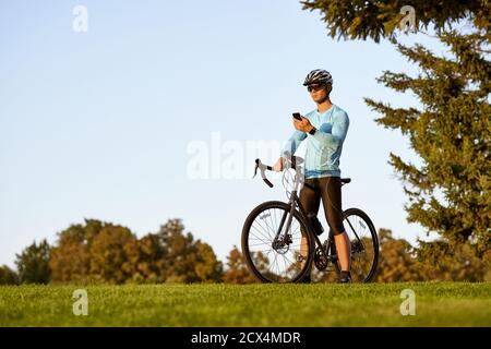 Faire une pause. Jeune homme sportif, cycliste professionnel en vêtements de sport et casque de protection debout avec son vélo dans le parc, en utilisant son smartphone et en vérifiant les résultats de fitness. Mode de vie actif Banque D'Images