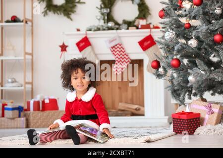 Adorable fille afro-américaine dans le livre de lecture de costume de Santa à la maison La veille de Noël Banque D'Images