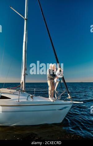 Amoureux de la mer. Pleine longueur de beau couple senior debout sur le côté de bateau à voile ou pont de yacht flottant dans la mer bleu calme, embrassant Banque D'Images