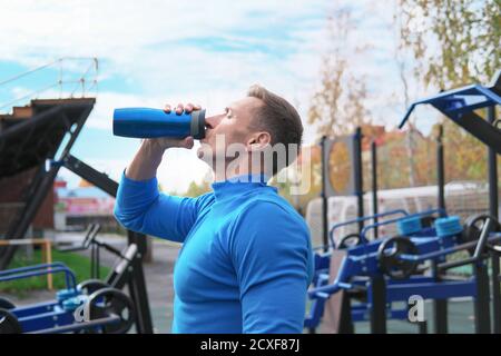 un homme musclé qui boit un cocktail de protéines de lactosérum après un entraînement sur le terrain de sport en plein air. stade d'automne, entraînement en plein air. sportif avec une bouteille de boisson sportive. Banque D'Images