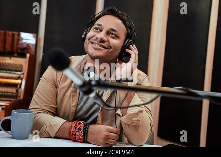 Jeune hôte de radio homme heureux regardant de côté pendant la diffusion en studio, à l'aide d'un microphone et d'un casque Banque D'Images