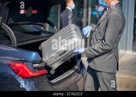 Homme élégant mettant une valise à l'intérieur du tronc pendant une pandémie Banque D'Images