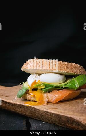 Bagel au saumon salé, aux épinards, à l'avocat et à l'œuf dur avec du jaune d'œuf liquide sur une planche à découper en bois sur fond noir de bois ancien. Banque D'Images