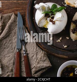 Burrata de fromage italien en tranches servi sur une planche à découper ronde avec des pignons, du pain en tranches, du basilic et des tomates séchées dans de l'huile d'olive avec fourchette et couteau o Banque D'Images