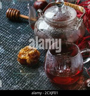 Thé à l'hibiscus rouge d'automne dans une tasse en verre et théière avec eau goutte à goutte, debout avec nid d'abeille sur fond de texture humide sombre avec feuilles d'érable d'automne et Banque D'Images