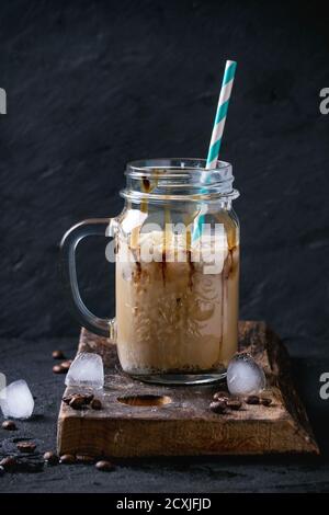Pot de maçon en verre de café glacé avec crème glacée et sauce au chocolat, servi avec des grains de café et des glaçons sur une planche à découper en bois sur fond noir texturé Banque D'Images