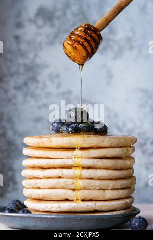 Crêpes aux myrtilles fraîches dans une assiette en céramique bleue, et miel provenant d'un pot de miel en bois, servies sur une nappe blanche avec un fond texturé bleu Banque D'Images