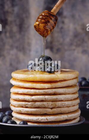 Crêpes aux myrtilles fraîches dans une assiette en céramique bleue, et miel fluide provenant d'un balancier de miel en bois, servies sur une surface en fer noir avec dos texturé d'étain Banque D'Images