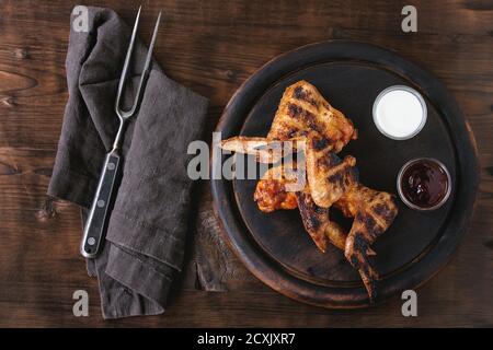 Ailes de poulet grillées au barbecue servies sur une planche à découper ronde en bois avec deux sauces, sel et fourchette de viande vintage sur du textile gris sur un dos en bois sombre Banque D'Images