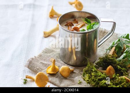 Soupe à la crème de chanterelles en fer tasse avec persil, servi avec des champignons frais et plus de mousse de forêt sur chiffon blanc éclatant.La lumière naturelle du jour. Style rustique. Banque D'Images