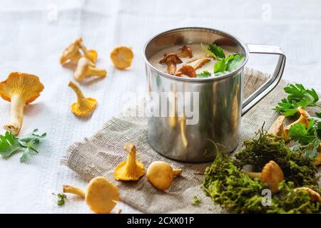 Soupe à la crème de chanterelles en fer tasse avec persil, servi avec des champignons frais et plus de mousse de forêt sur chiffon blanc éclatant.La lumière naturelle du jour. Style rustique. Banque D'Images