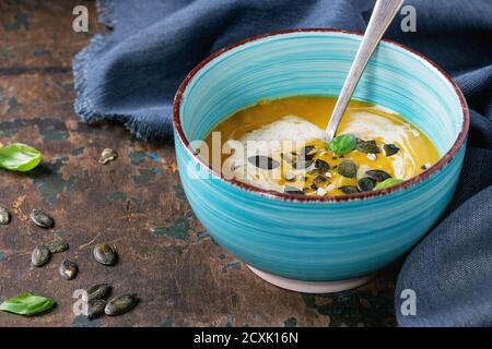 Bol en céramique turquoise de potiron et soupe douce à la crème de pommes de terre avec basilic frais, crème et graines, servi avec une cuillère sur une serviette en textile sur du vieux bois de tex Banque D'Images